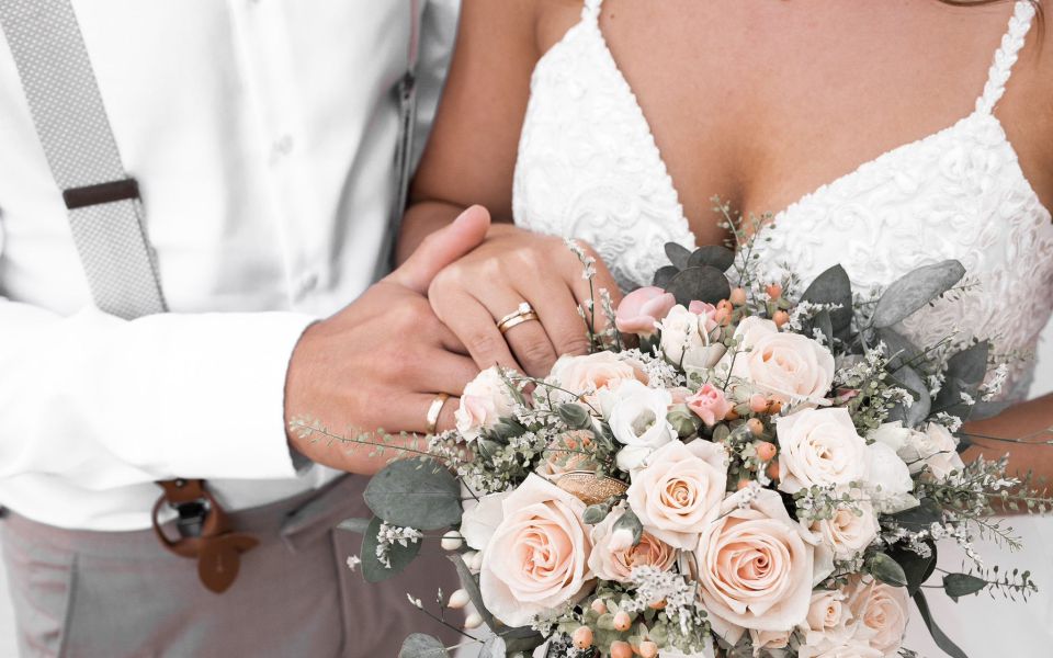 Die Hände eines Brautpaares mit Eheringen und deren Brautstrauß.